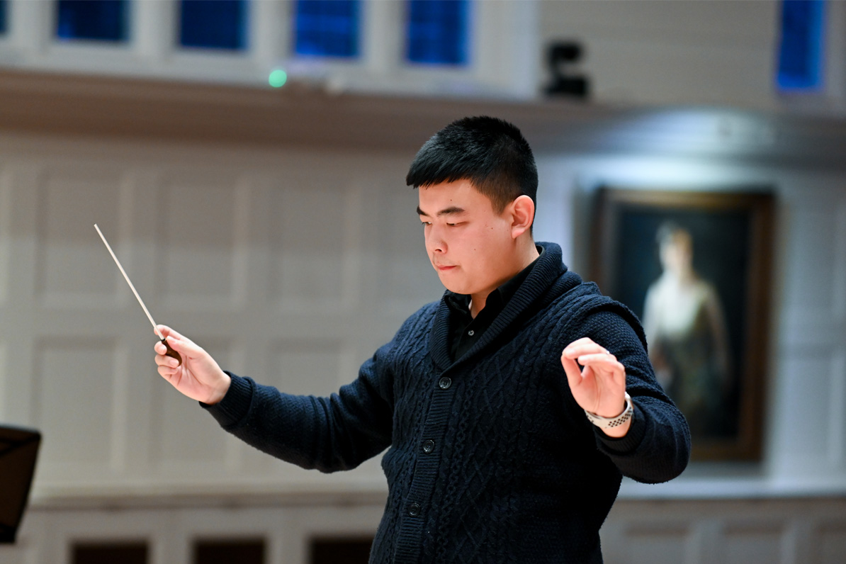 A male student, wearing a smart black shirt, conducting a brass ensemble in a performance room.